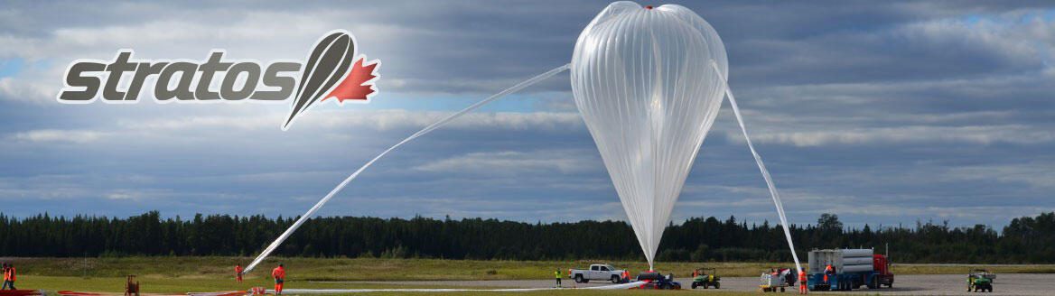A large stratospheric helium balloon partially inflated on an airfield, held by ground crew in high-visibility jackets, under the Stratos programme banner.