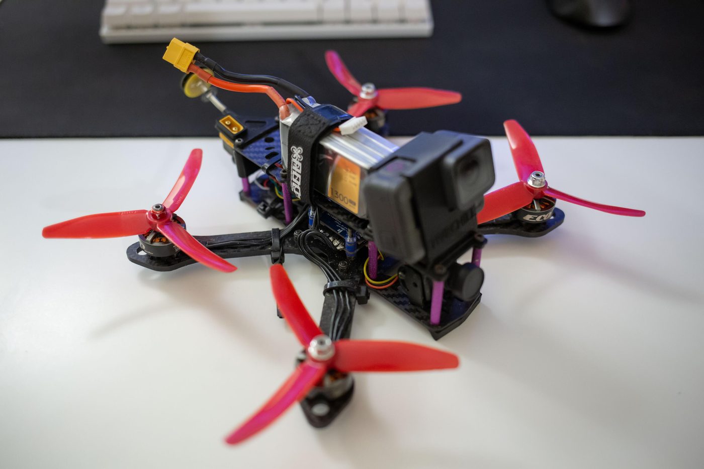 2020 FPV quadcopter build on a desk, with red three-blade propellers, a carbon-fiber frame, strapped LiPo battery and a GoPro mounted on the nose.