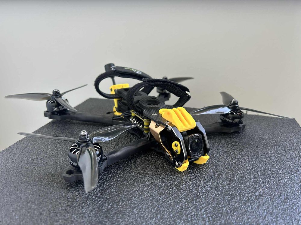 2025 FPV quadcopter build with black propellers, a yellow 3D-printed camera housing and a DJI O3 camera at the nose.